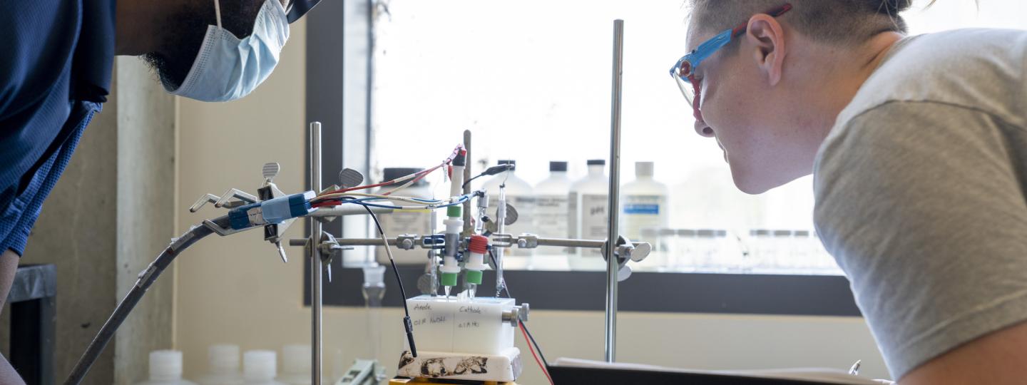 Two students wearing lab glasses observe an experiment featuring an anode and a cathode hooked up to various wires.
