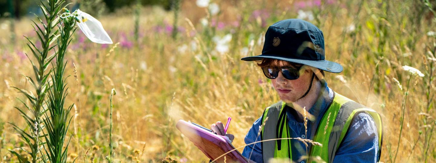 A student crouches in a field holding a clipboard.