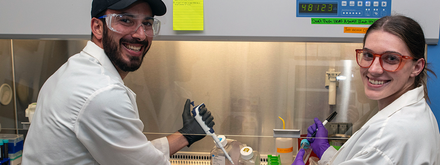 Two people in lab coats and safety glasses with pipettes in their hands stand in front of a fume hood in a laboratory smiling.