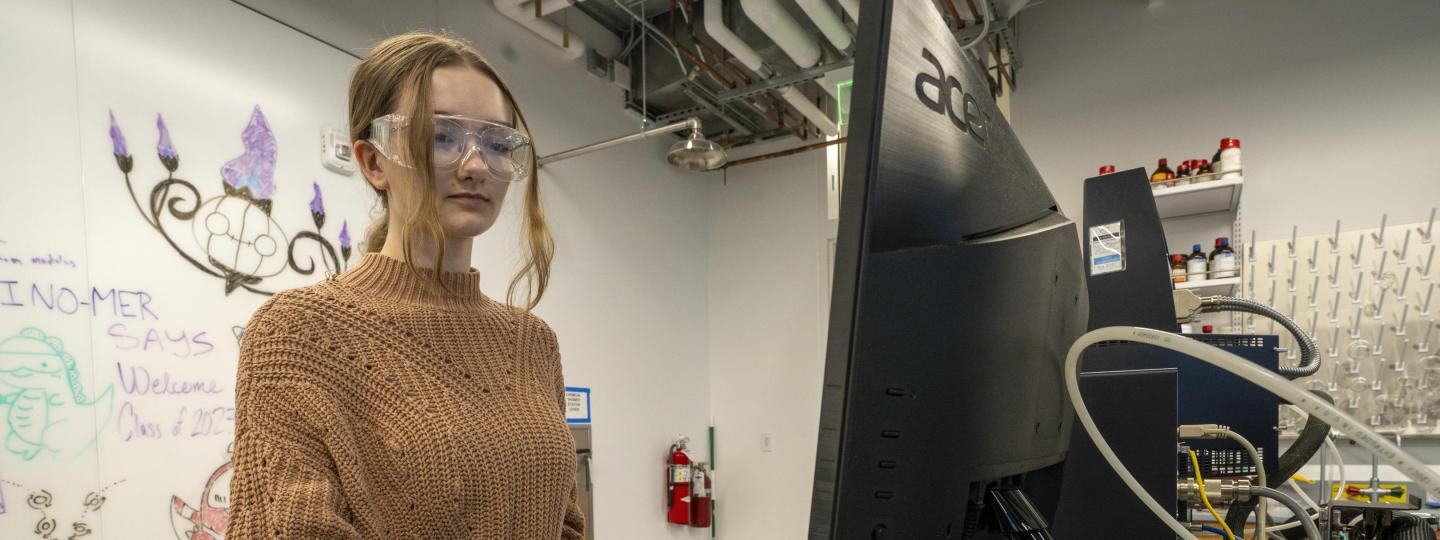 A student wearing gloves and safety glasses uses a computer in a lab.