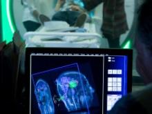 In the foreground a monitor displays a scan of a person's head. In the background, a person is seen in an MRI machine.