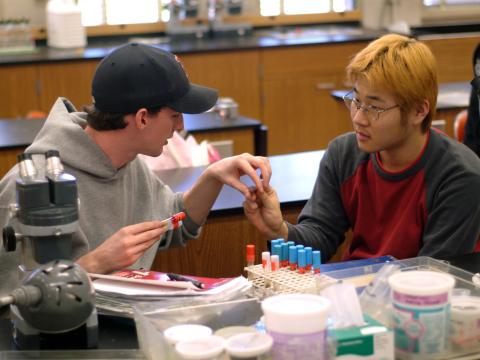 A person hands another person a test tube while sitting together at a table covered with equipment and a microscope in a lab.