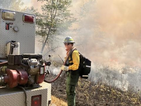 Meredith Jacobson poses behind a firetruck in wildland firefighter gear. A fire and smoke from a prescribed burn is visible in the background.