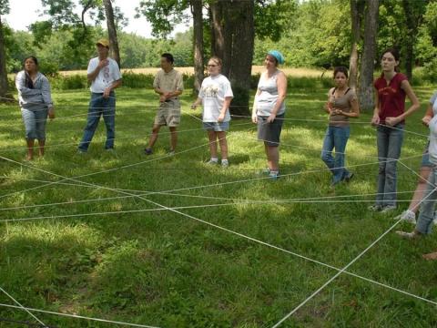 A group of teens and adults stand in a circle, each holding the edges of a string that is crossed to form a messy web between them.