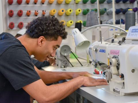 A student works at a sewing machine. Brightly colored spools of thread line the wall behind the student