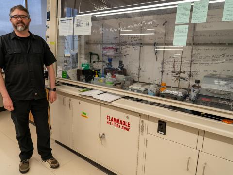 A portrait of Darren Johnson wearing lab glasses standing beside a fume hood in a lab. The fume hood is covered in chemical equations written on the glass.