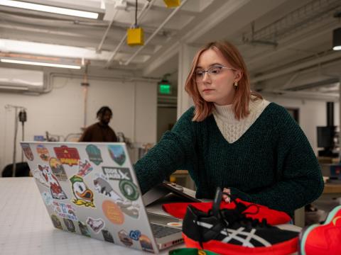 A student sitting at a table with a laptop computer and several athletic shoes.