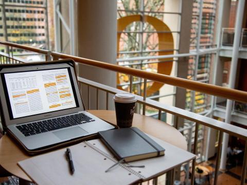 A table with an open laptop, notebook, and cup of coffee.