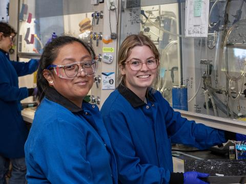 Two people in lab coats and safety glasses smile while standing in a laboratory.