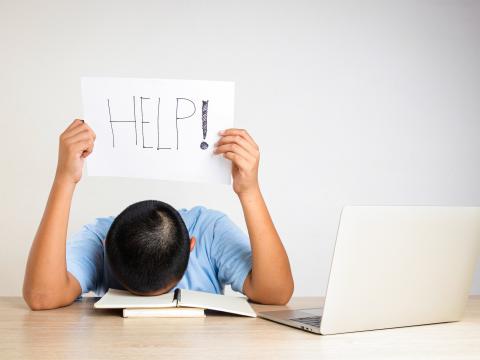 A student with their head on a desk holds up a sign reading "help."