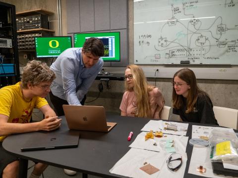 A group of students look at a laptop as a faculty member points to something on the screen.