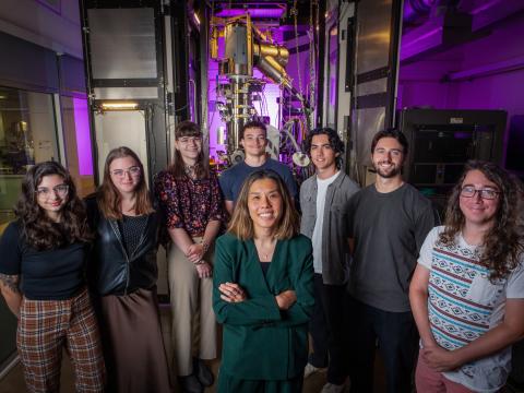 A group of young scientists stands in front of a scanning transmission electron microscope in a low-lit room.