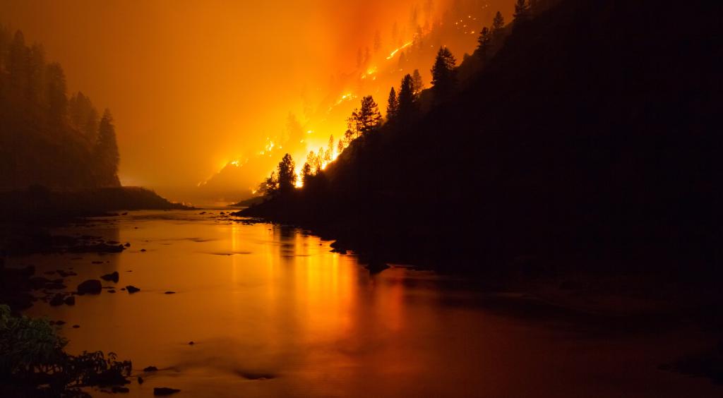 A fire burns a hillside at night beside a river, off the surface of which the light of the fire is reflected.
