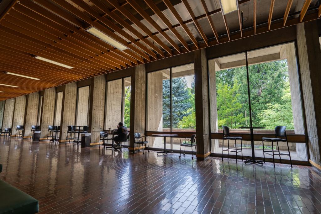 A person sits at a table in front of large floor-to-ceiling windows in McKenzie Hall on the UO campus.