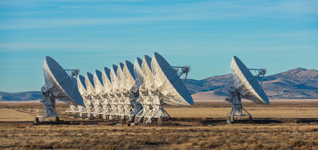 A long row of large white radio telescopes stands in an open desert plain under a blue sky.