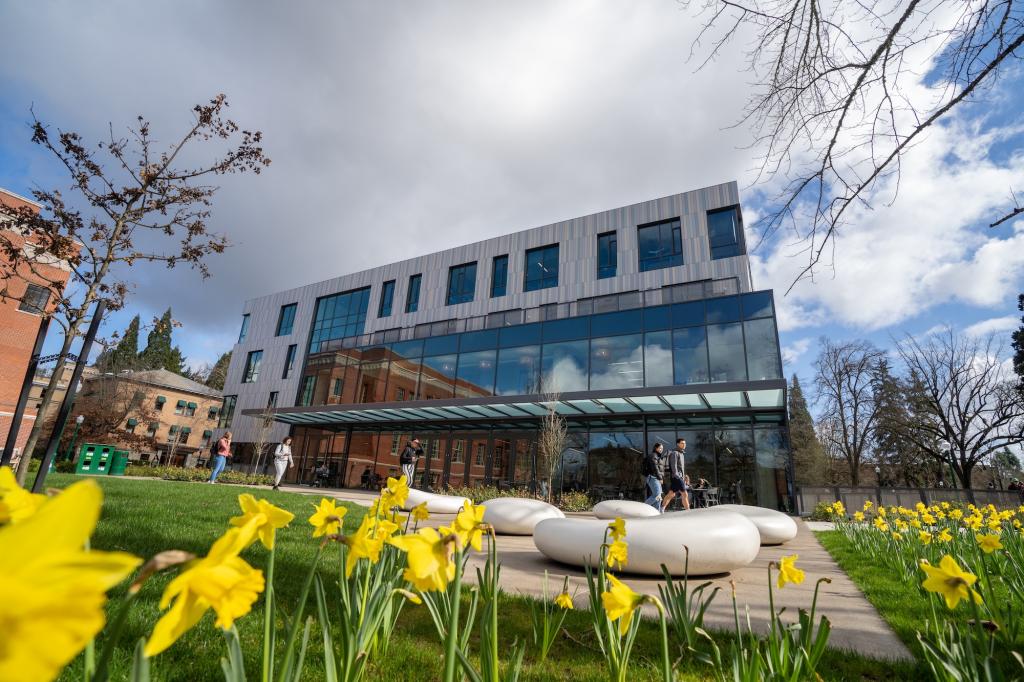 A modern multi-story glass and metal building on a college campus, with bright yellow flowers in the foreground.