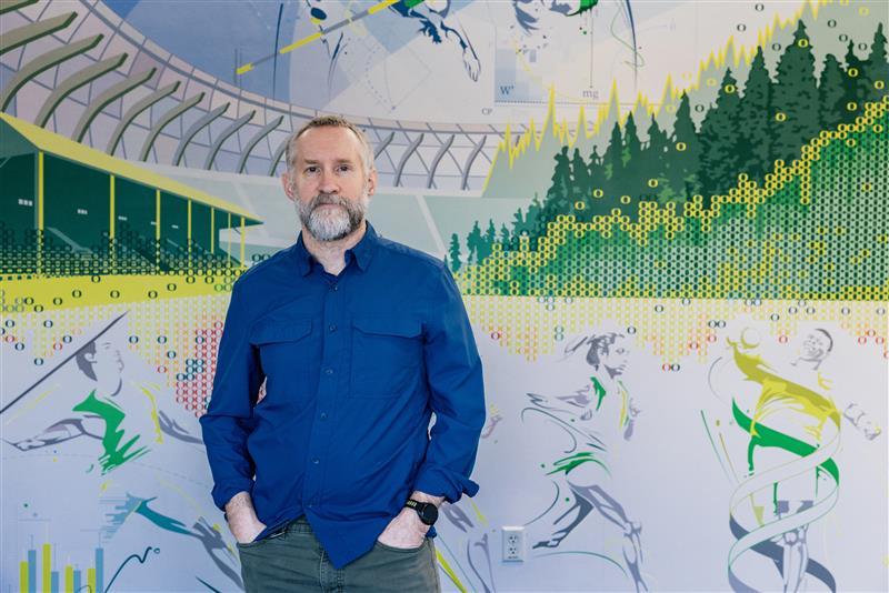 A portrait of a man, John Halliwill, standing in front of a mural of Hayward Field.