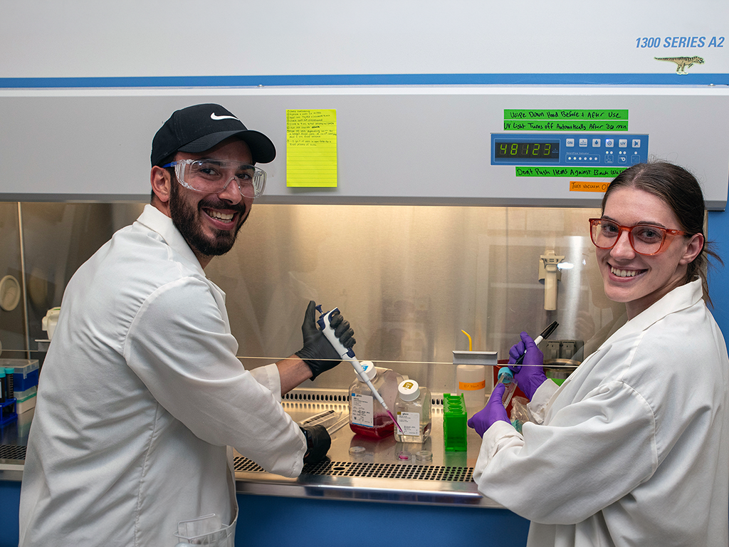 Two people in lab coats and safety glasses with pipettes in their hands stand in front of a fume hood in a laboratory smiling.