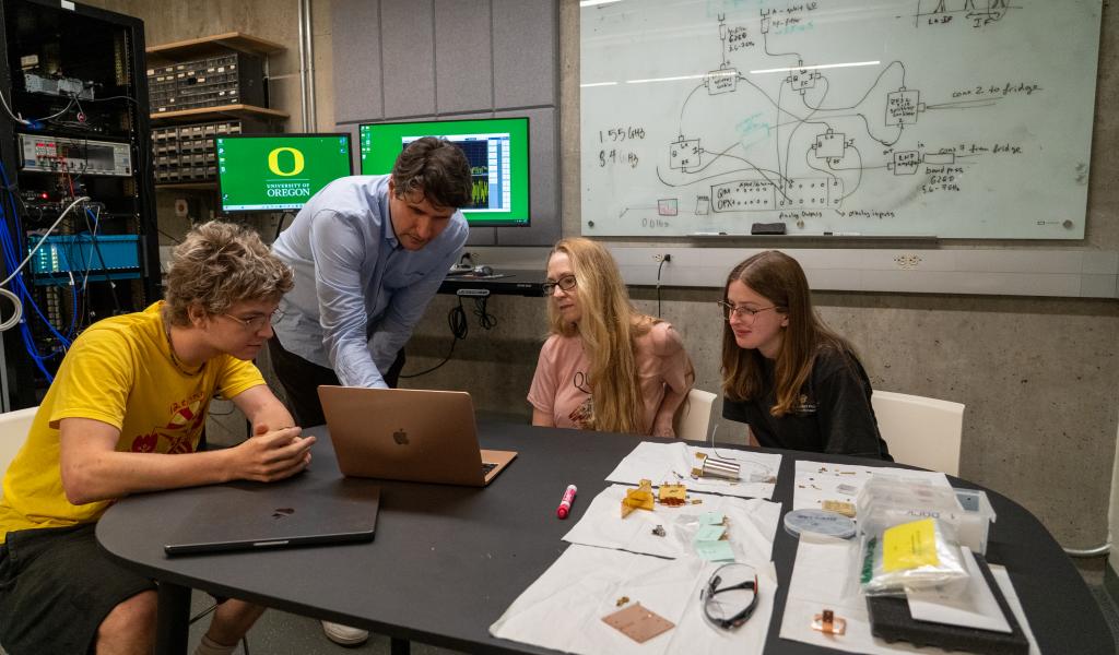 A group of students look at a laptop as a faculty member points to something on the screen.