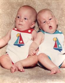 Two infants in identical outfits are positioned on a blanket for a portrait.