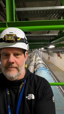 A man wearing a hardhat stands in the Large Hadron Collider underground accelerator tunnel in which several pipes disappear into the distance.