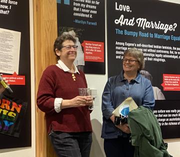 Two women stand against a wall covered in posters from the Outliers and Outlaws exhibit, both smiling.