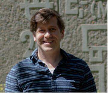 A portrait of Ben McMorran, who is wearing a blue shirt, smiling, and standing in front of a stone wall with various symbols from chemistry and physics etched onto it. 