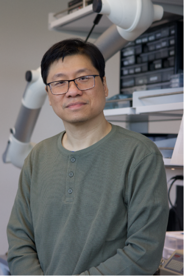 Man wearing glasses subtly smiling into the camera stands in front of a blurred lab setting