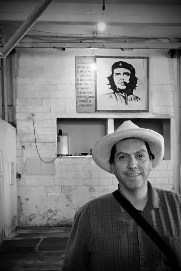 A man in a hat stands in front of a poster of Che Guevara.