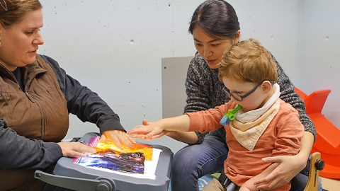 Two adults help a small child feel plastic bags full of different liquids as part of a sensory experience.
