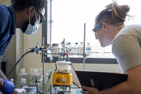 Two students wearing lab glasses observe an experiment featuring an anode and a cathode hooked up to various wires.