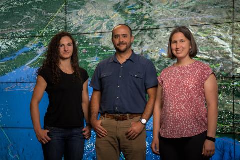 A portrait of Valerie Sahakian, Diego Melgar Moctezuma, and Amanda Thomas standing in front of a map of the US West Coast projected on a wall of monitors.