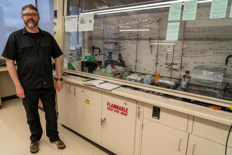 A portrait of Darren Johnson wearing lab glasses standing beside a fume hood in a lab. The fume hood is covered in chemical equations written on the glass.