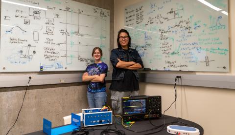 Two people stand with crossed arms in front of white boards covered in calculations by a table covered in electronics equipment.
