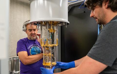 Two people talk to each other while the person in the foreground holds their hands around the base of a cryostat instrument.