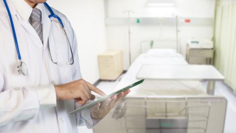 Man in lab coat with stethoscope and clipboard stands in front of hospital bed