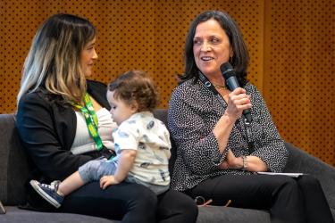 Two women sit in a chair. One holds a baby and the other holds a microphone.