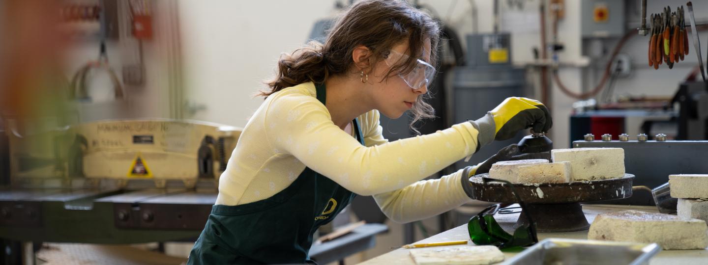 Woman wearing protective glasses and gloves leans over a table full of metal working supplies.
