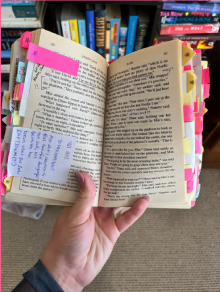 A person's hand holds a book that is heavily marked with sticky notes and underlined text.