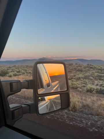 Sunset reflected in the rearview mirror of an F-250 pickup truck. Beyond the mirror, in the distance, a field of sagebrush and pink-tinted clouds above mountains.