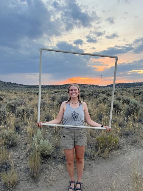 A portrait of Zoey Bailey, who is holding a square frame made of plastic PVC pipe, standing in front of a field of sagebrush at sunset.